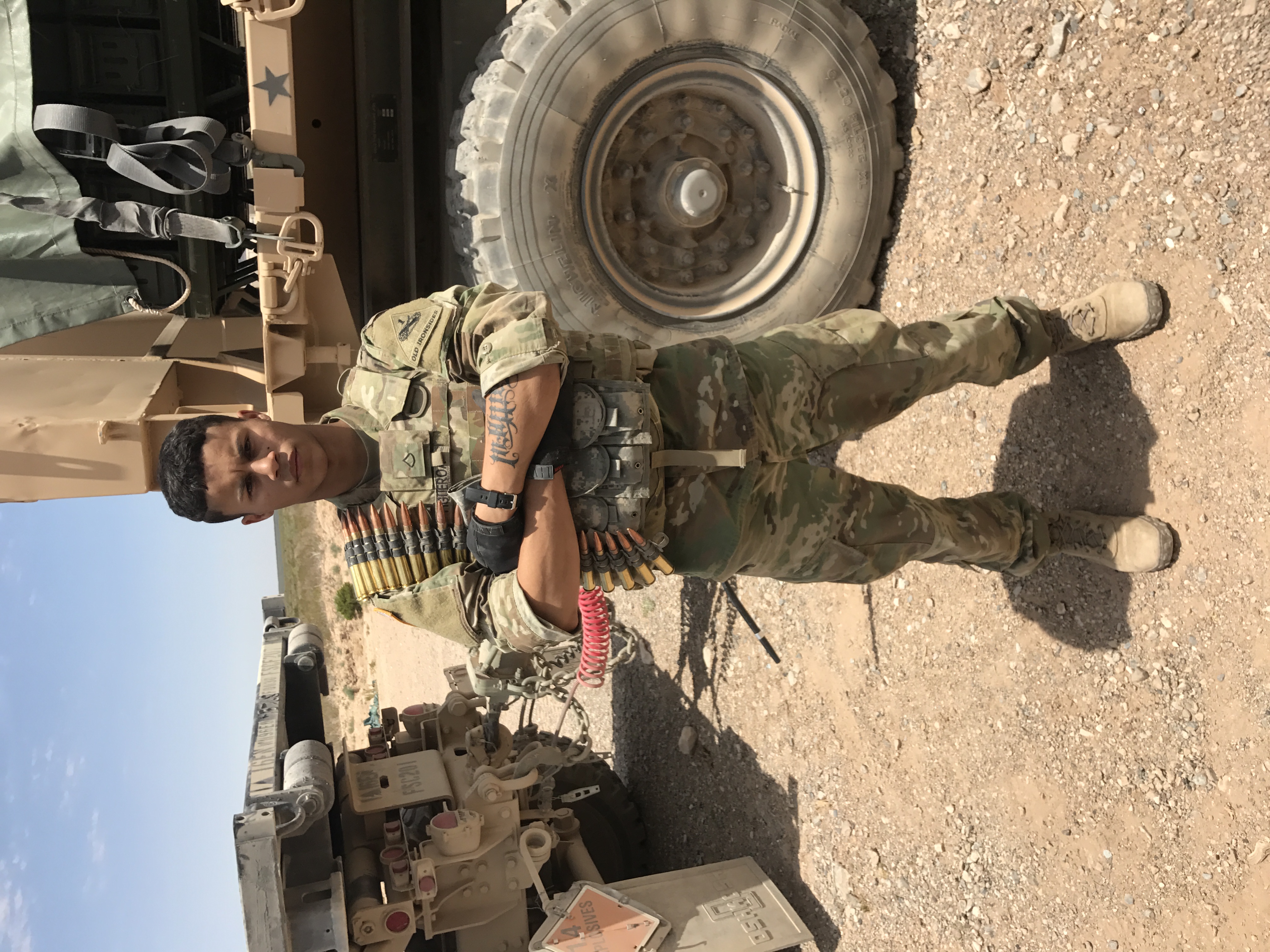 Ason in military uniform with tactical gear, standing in desert environment with military vehicles in background
